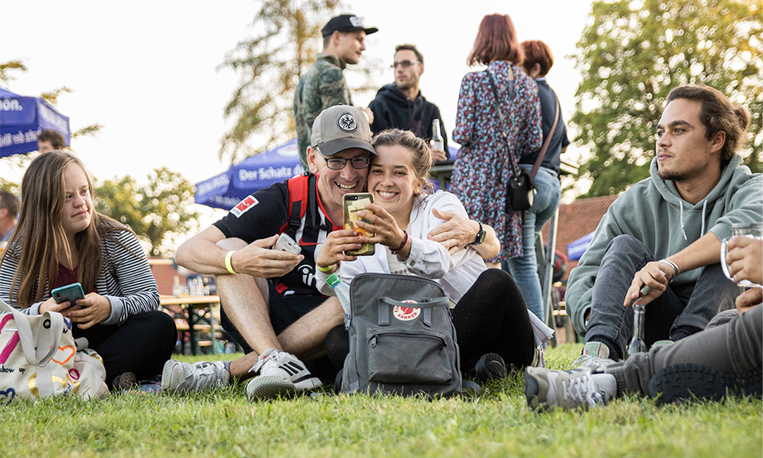 Menschen sitzen bei einem Sommerfest auf der Wiese, zwei Personen halten sich im Arm und machen mit dem Handy ein Selfie
