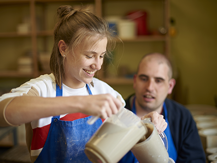 Eine junge lächelnde Frau beim Töpfern mit einem Mann mit Behinderung im Hintergrund